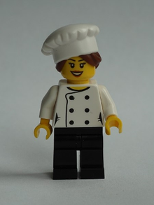 Minifig chef026 - Chef - Black Legs, Open Mouth Smile, Hair in Bun, 'LEGO HOUSE Home of the Brick' on Back, Female - De BrickPlaats